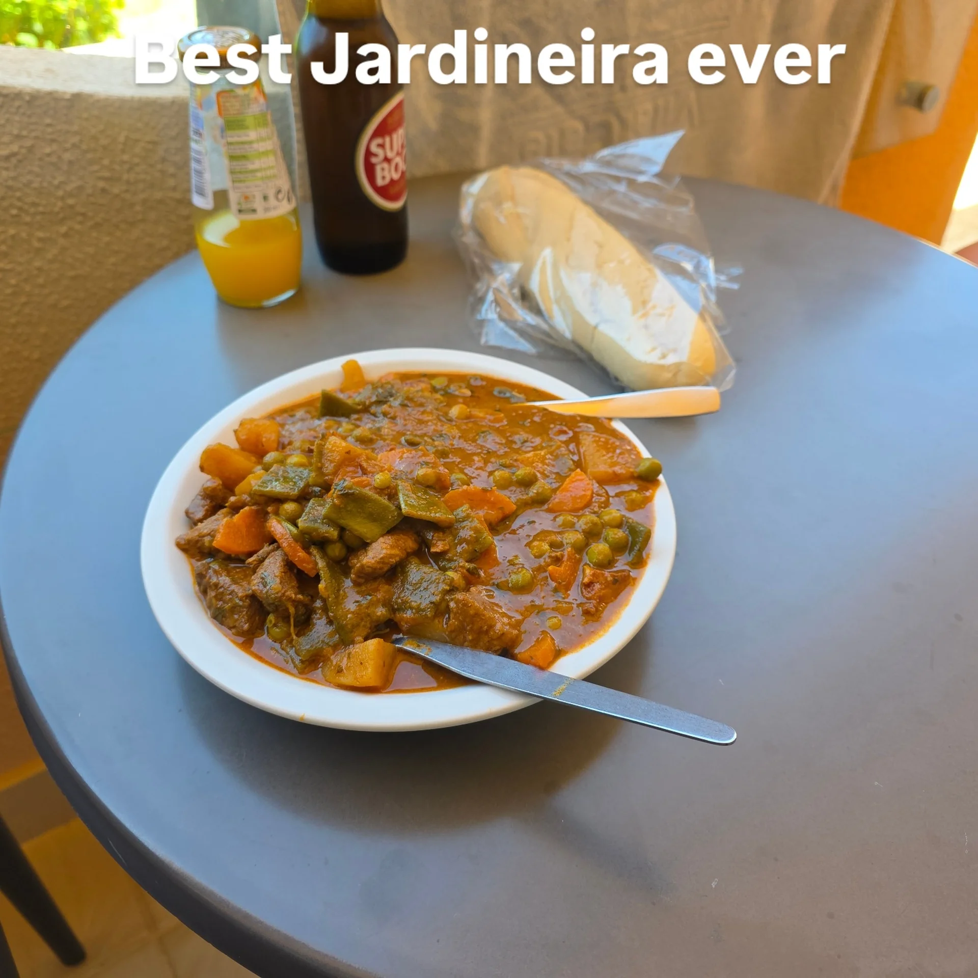 Ein Teller traditioneller portugiesischer Rindfleisch-Eintopf (Jardineira) mit Gemüse auf einem Balkontisch, flankiert von einer Flasche Super Bock Bier und Baguette.
