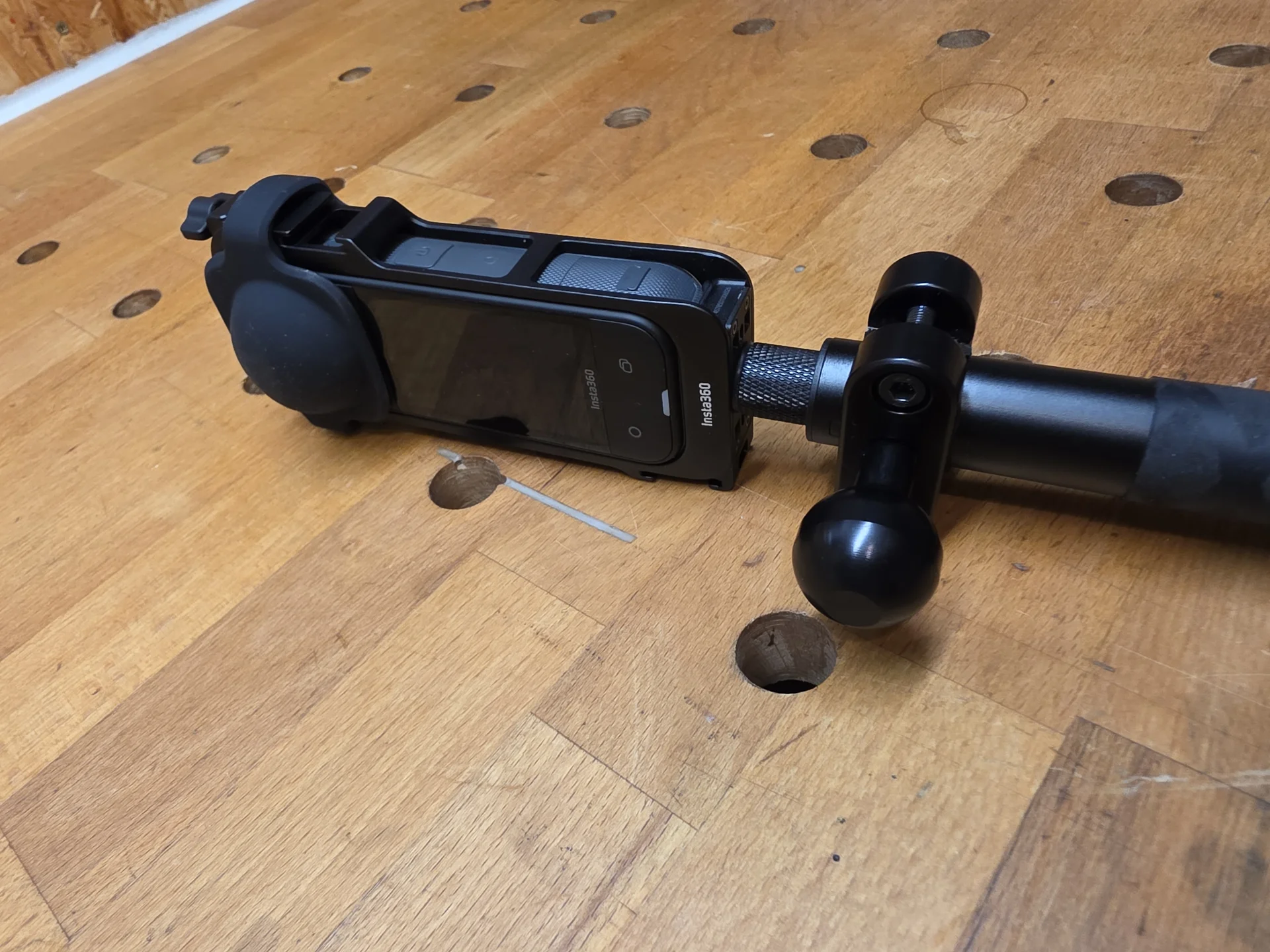 Seitliche Nahaufnahme einer Insta360 X5 mit Kugelflex-Klemmschelle am Teleskopstab auf einer Holzwerkbank.